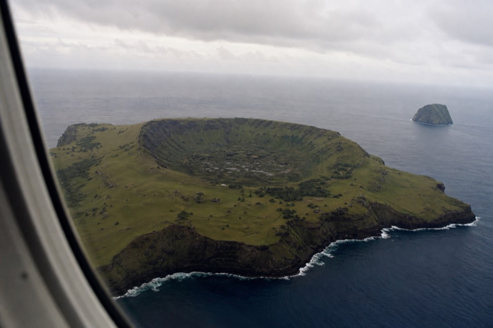 View from an aircraft window of a small green volcanic island surrounded by deep blue open ocean