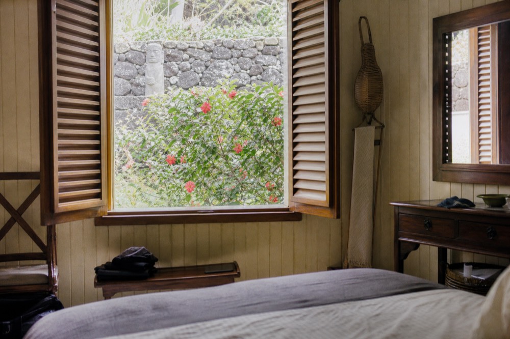Simple guesthouse room with wooden shutters open to a garden of hibiscus and volcanic stone wall