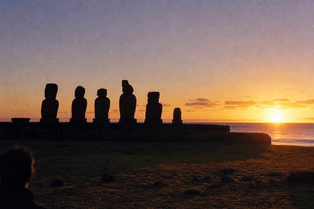 Row of moai in silhouette against an orange and violet dawn sky with low cloud on the horizon