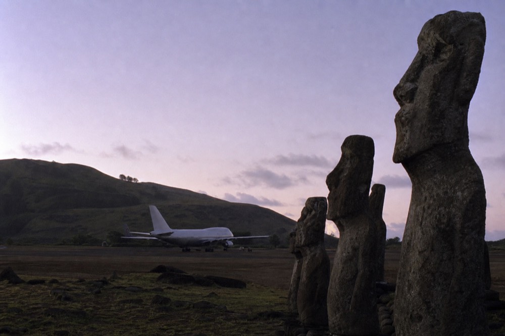 Wide-body aircraft parked on the single apron at Mataveri airfield with green volcanic hills rising behind the runway