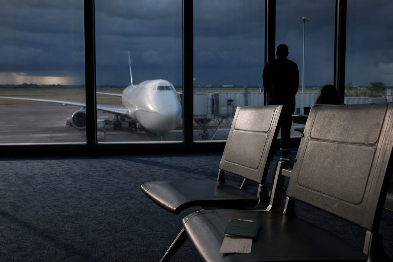 Passport and boarding pass lying on a departure-gate seat with a wide-body aircraft visible through the terminal glass beyond