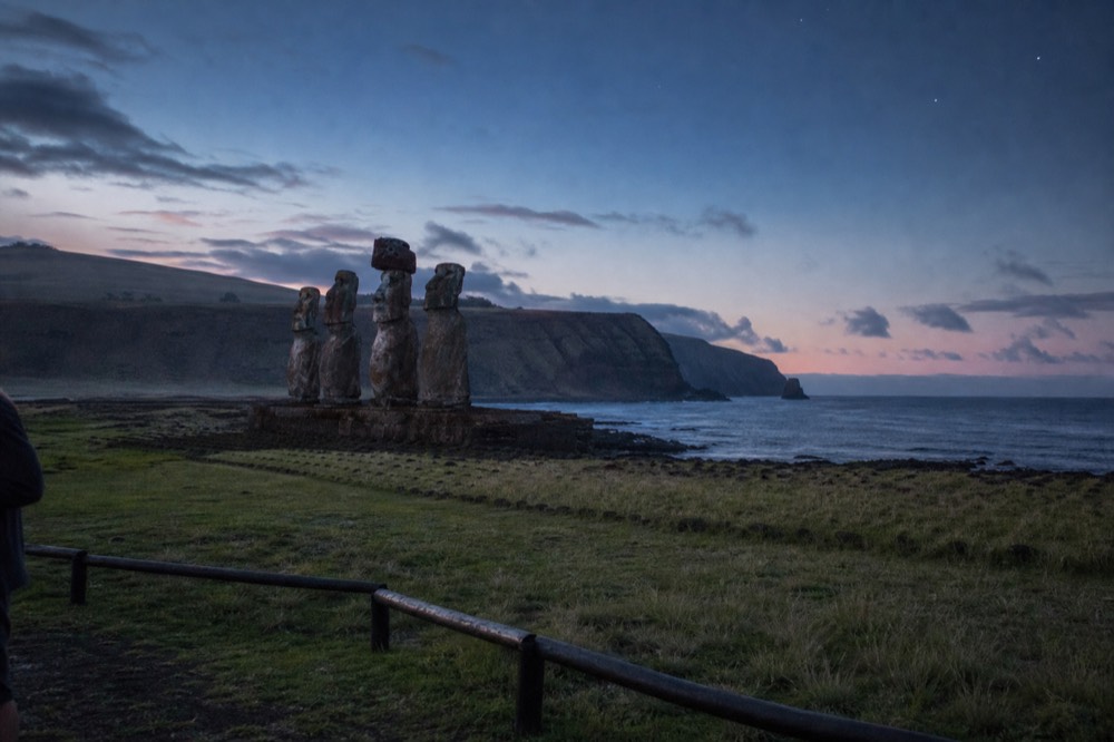 Weathered moai on a stone platform beside the ocean with fresh grass and a low fence keeping visitors back