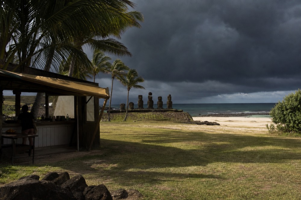 Food kiosk with a shaded awning at the edge of the sand at Anakena, palms leaning over pale coral beach beyond