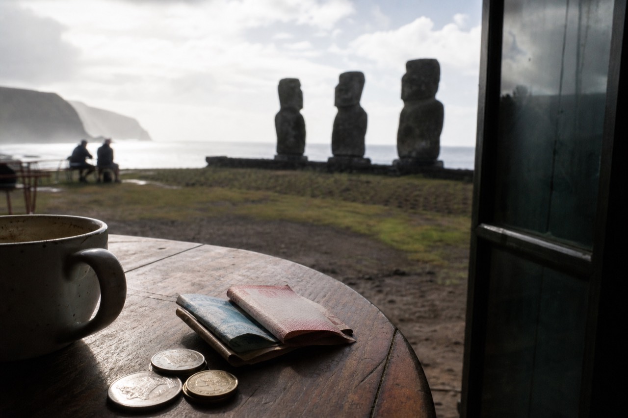 Folded Chilean peso notes and coins on a wooden cafe table beside a coffee cup with the harbour visible through an open doorway