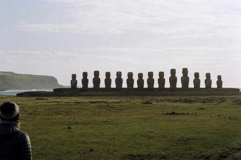 Fifteen moai standing in a row on a long restored stone platform silhouetted against a pale morning sky