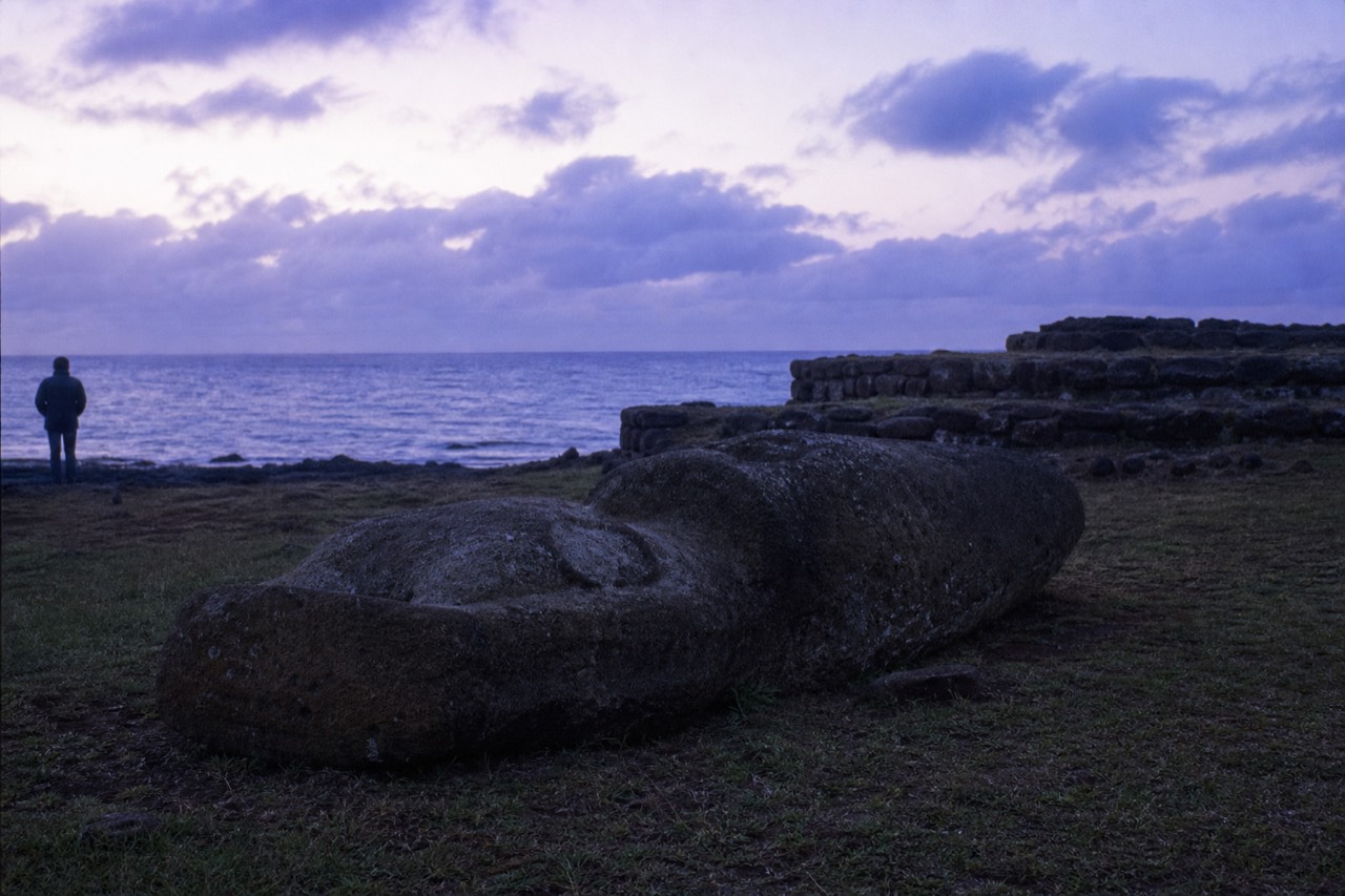 Toppled moai lying face down in coastal grass beside a ruined stone platform with the Pacific behind
