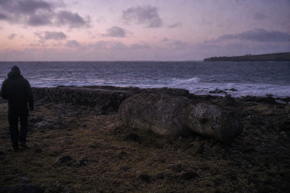 Toppled moai lying face down beside a low stone platform on a rocky shoreline with waves breaking behind