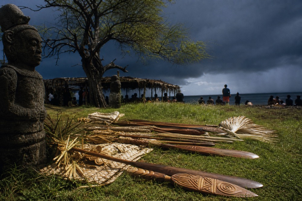Carved wooden ceremonial paddles and woven palm decorations laid out on grass before a village gathering