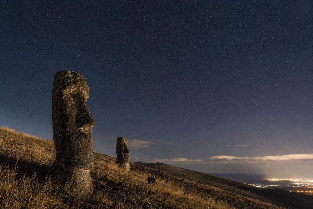 Grassy volcanic ridge at night with a faint town glow low on one horizon and stars filling the rest of the sky