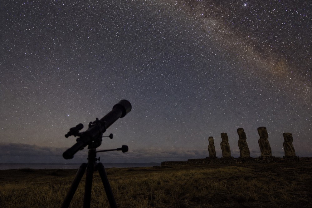 Telescope tube silhouetted against a dense field of southern stars above a dark grassy horizon