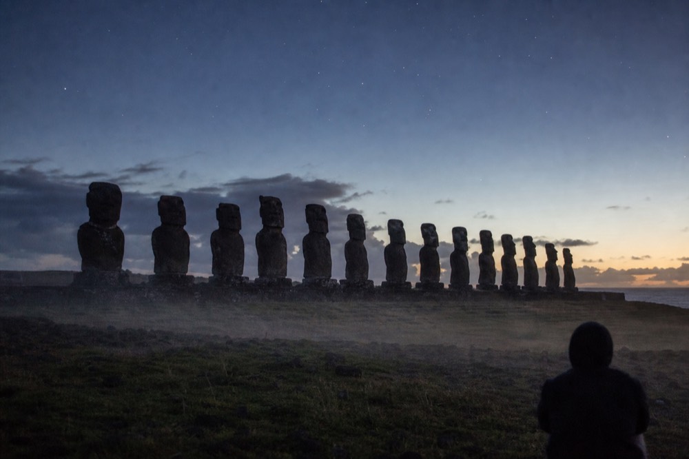 Row of moai in near-darkness against a faintly lightening sky with stars still visible above the platform