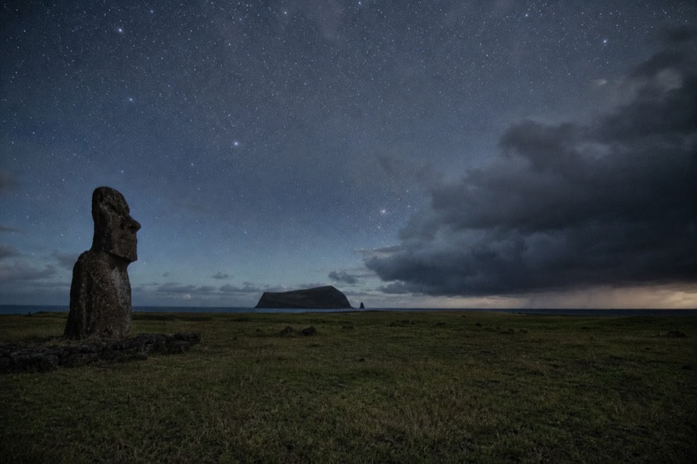 Wide field of southern stars above an empty grass ridge with a single distant headland in silhouette