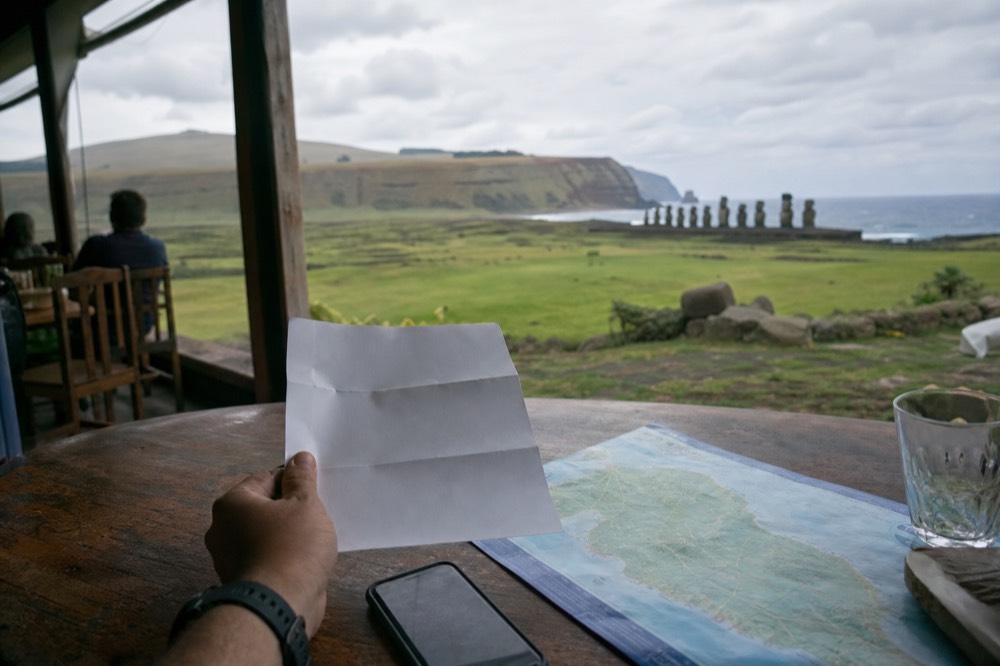 Hand holding a printed itinerary sheet on a cafe table with a phone and a map of the island beside it