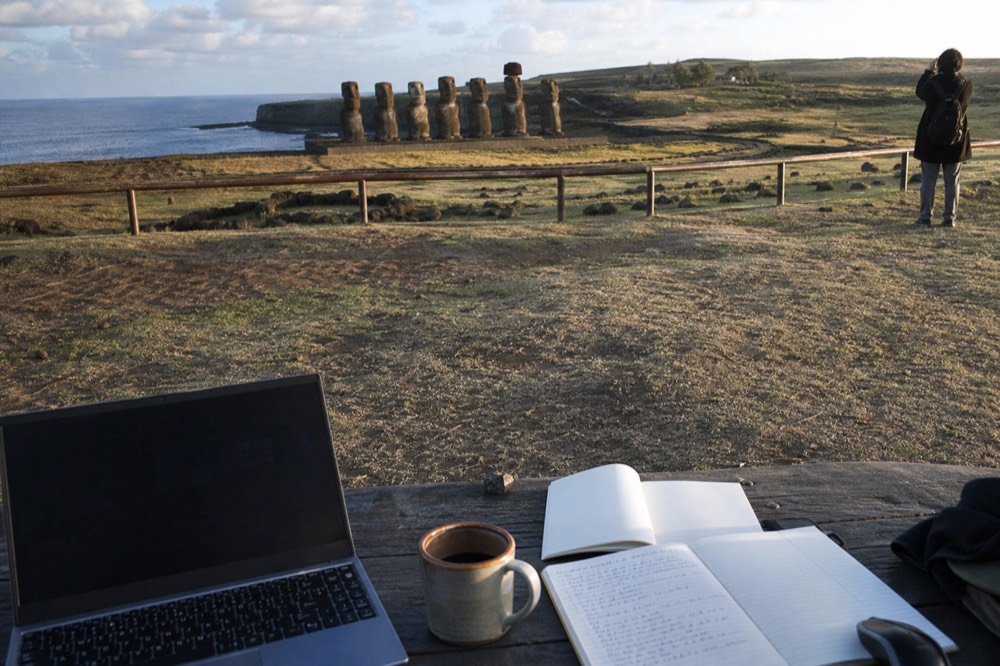 Two open notebooks with handwritten cost columns beside a laptop and a cup of coffee on a wooden table