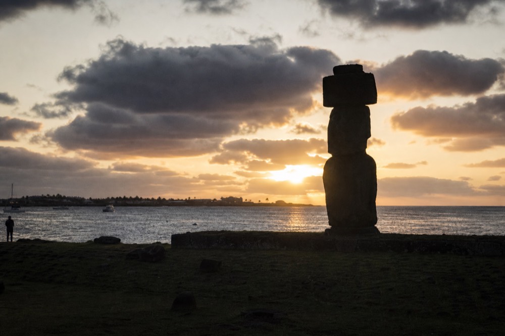 Sunset silhouette of a single moai at the Tahai complex with the harbour and low cloud behind