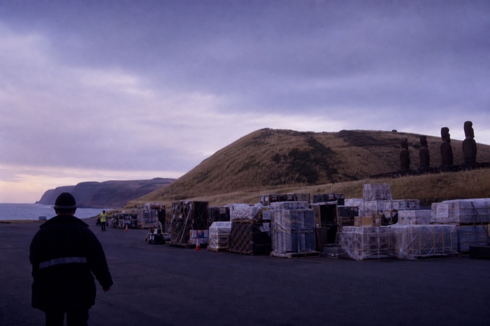 Cargo pallets and crates stacked on an airport apron with a hillside of dry grass behind them