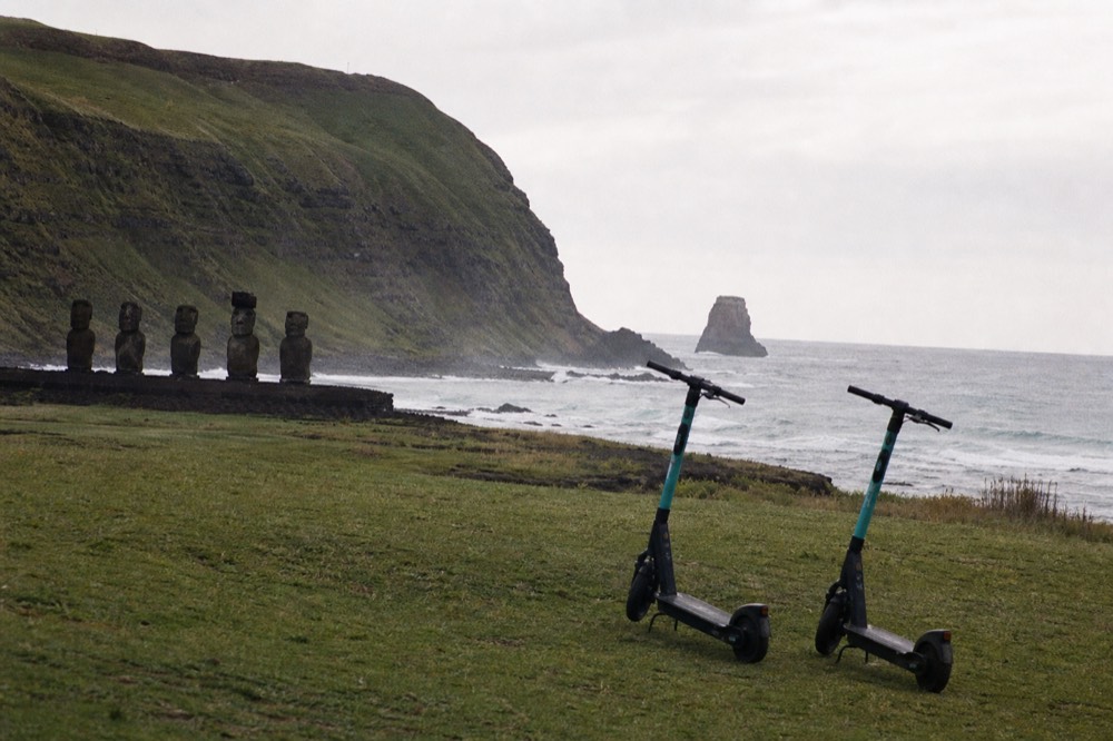 Two rental scooters parked on a grass verge above a rocky coastline with surf breaking on black rock