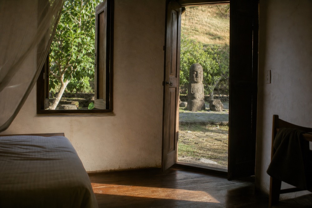 Simple guesthouse room with white walls, a wooden bed frame and open shutters looking onto a garden