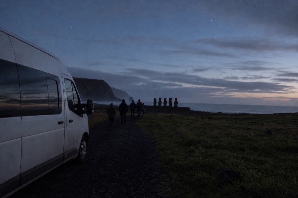 Tour van parked on a gravel track with a small group walking toward a coastal stone platform in the distance