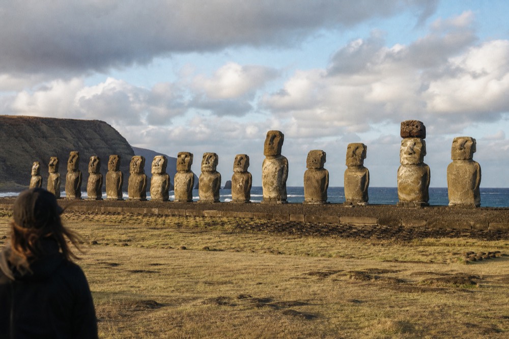 Line of fifteen restored moai standing on a long stone platform under early cloud with the coastline behind
