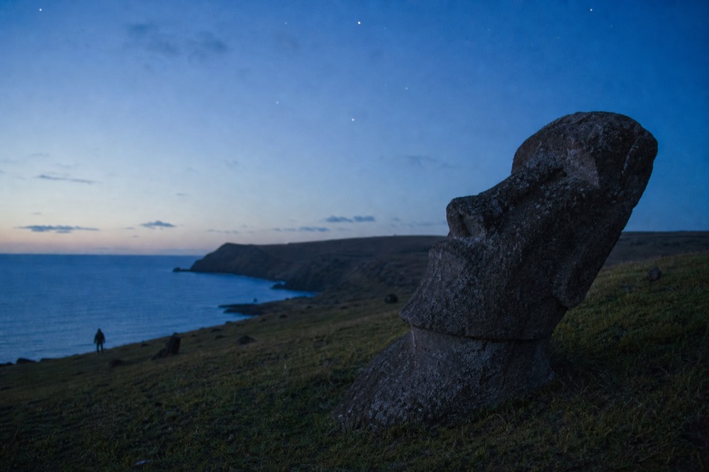 Weathered stone moai torso half sunk in green grass on a volcanic hillside, its face tilted toward the sky