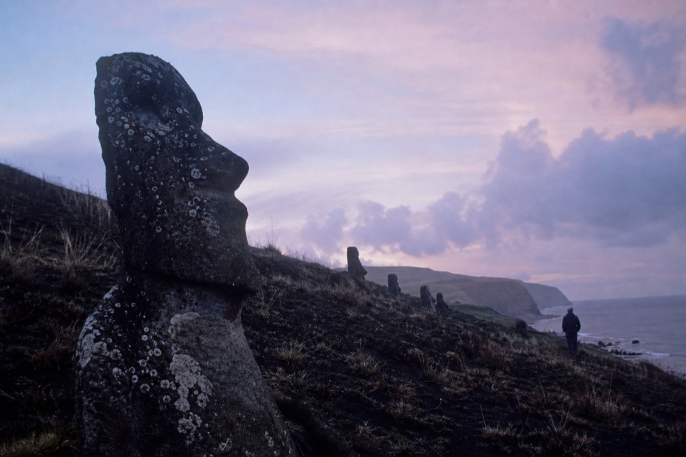 Burnt grass and darkened volcanic stone on a quarry hillside with a scorched moai shoulder in the foreground