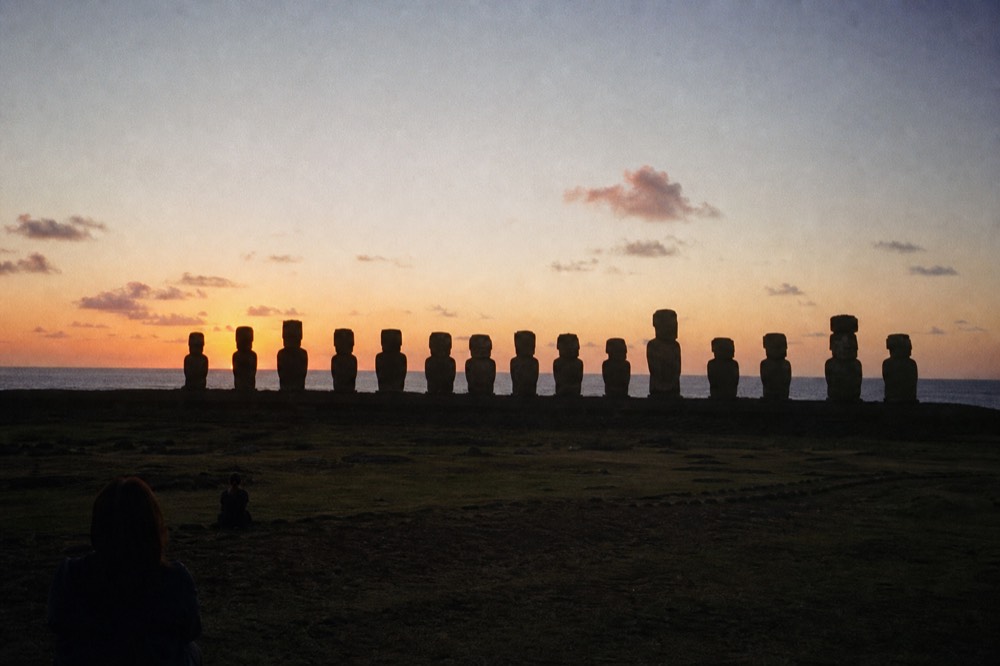 Fifteen moai in silhouette on a long platform against a pale orange pre-dawn sky above the Pacific