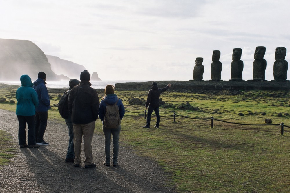 Small group standing on a marked gravel path listening to a guide with a stone platform and moai beyond the rope line